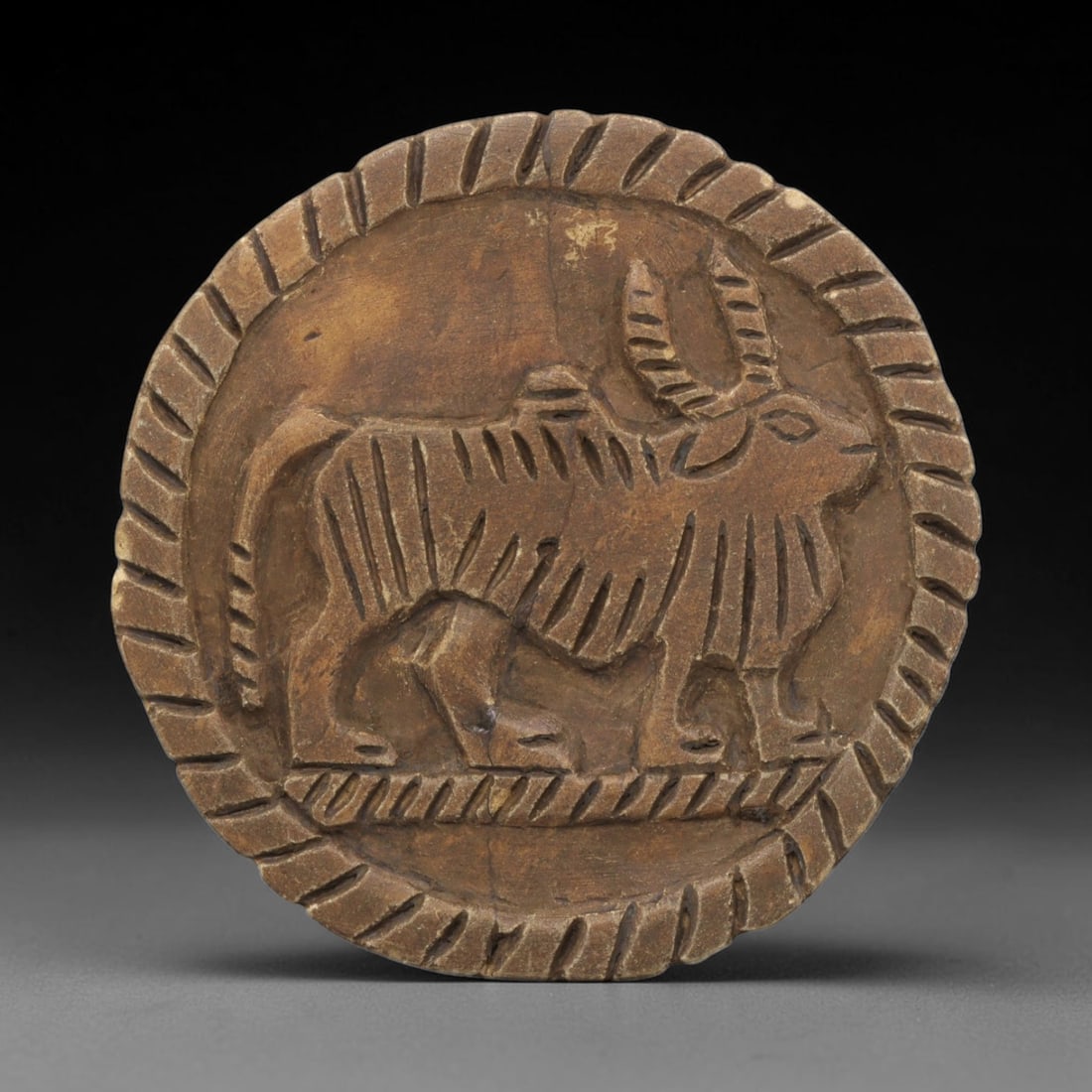 Ancient Steatite Plaque - Near Eastern Bull Relief, 22ZY: Carved from steatite, this circular plaque features a relief depiction of a bull, rendered in the schematic artistic tradition of the Ancient Near East. The bovine figure is shown standing in profile,