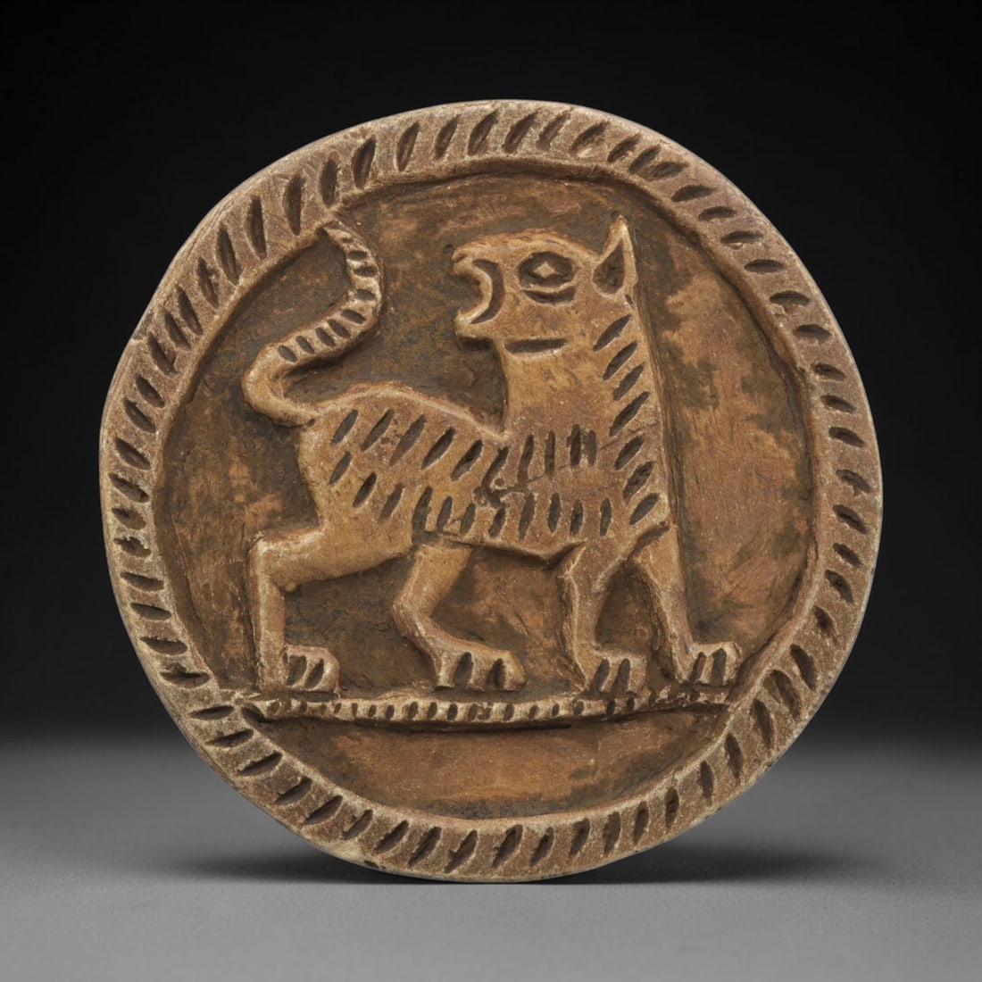 Ancient Steatite Plaque - Near Eastern Lion Relief, 27ZY: Carved from steatite, this circular plaque features a relief depiction of a stylized lion, rendered in the schematic artistic tradition of the Ancient Near East. The feline figure is shown standing in