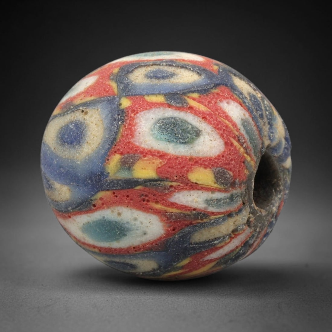 Ancient Mosaic Glass Bead - Roman Eye Pattern Amulet, 51WD: This spherical bead is crafted from core-formed mosaic glass, featuring a vibrant red base decorated with intricate concentric eye motifs. Each motif consists of a central glass circle in shades of bl
