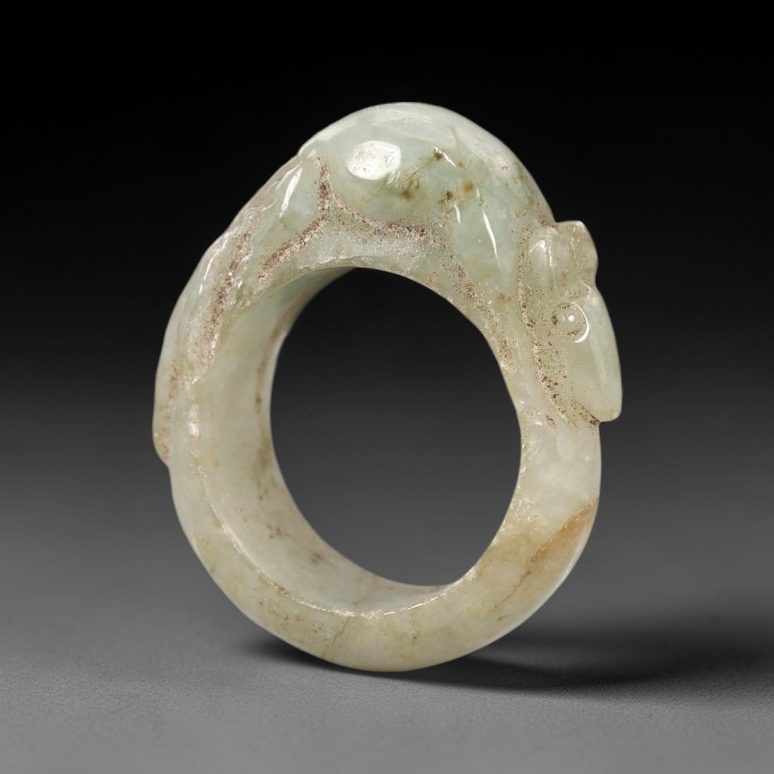 A Chinese Jadeite Jade Mouse-Form Ring, Qing Dynasty, 63FS: Carved from pale greenish-white jadeite jade with natural brownish inclusions and subtle internal veining, the object is formed as a circular ring with a raised, carved mouse integrated into the upper