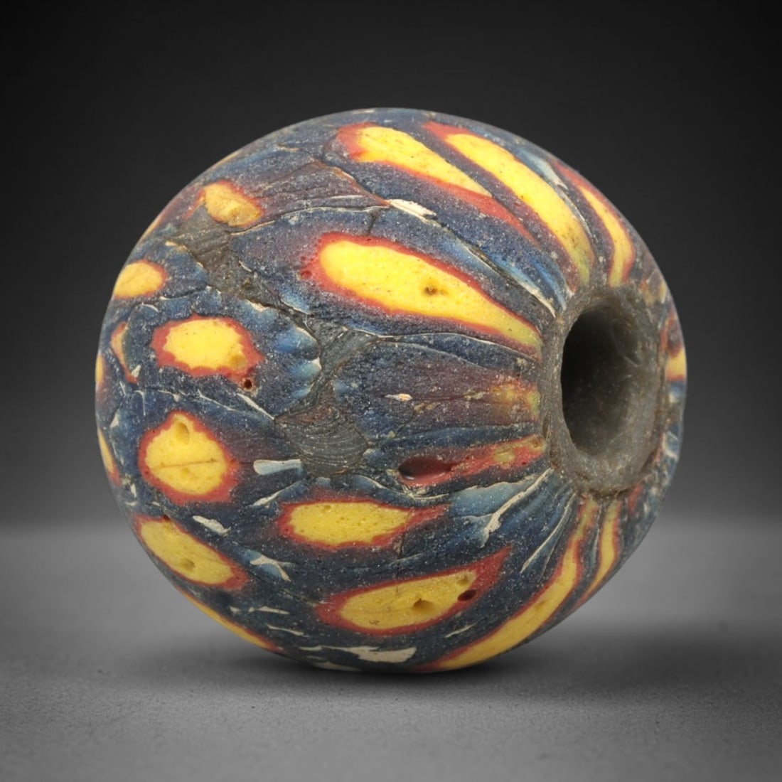 Ancient Mosaic Glass Bead - Roman Swirl Pattern Amulet, 66WD: This spherical bead is crafted from core-formed mosaic glass, featuring a deep indigo blue base decorated with dynamic swirling motifs in bright yellow, vivid red, and opaque white glass canes. The de