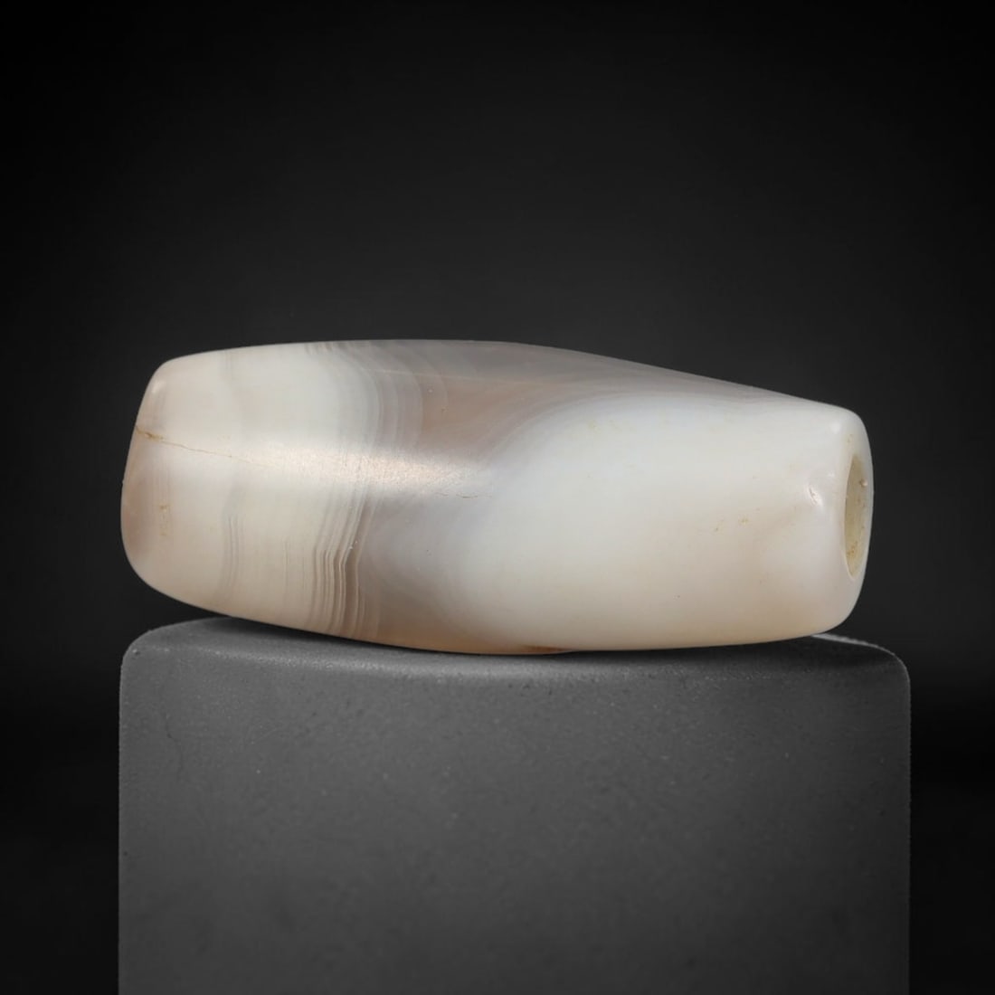 Ancient Banded Agate Bead - Near Eastern Artifact, 41BD: This artifact is crafted from natural banded agate, shaped into an elongated barrel form with fine, wavy concentric banding in soft cream and taupe hues. The bead features a central perforation for su
