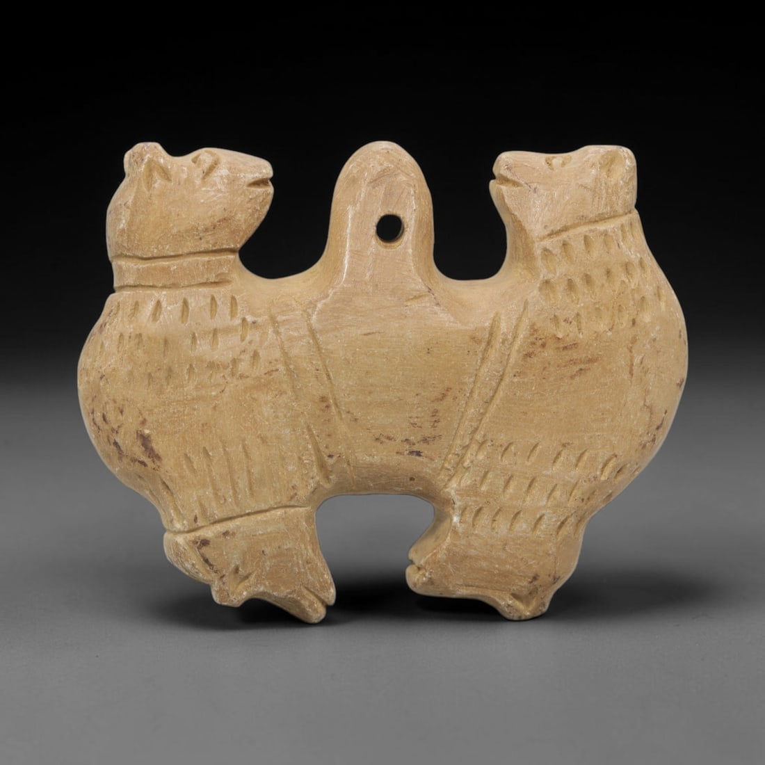 Ancient Stone Double Zoomorphic Amulet - Near Eastern, 17WJ: Carved from a fine-grained, pale beige stone, this artifact takes the form of a symmetrical double zoomorphic amulet, depicting two confronting animal figures joined at a central suspension loop. Each