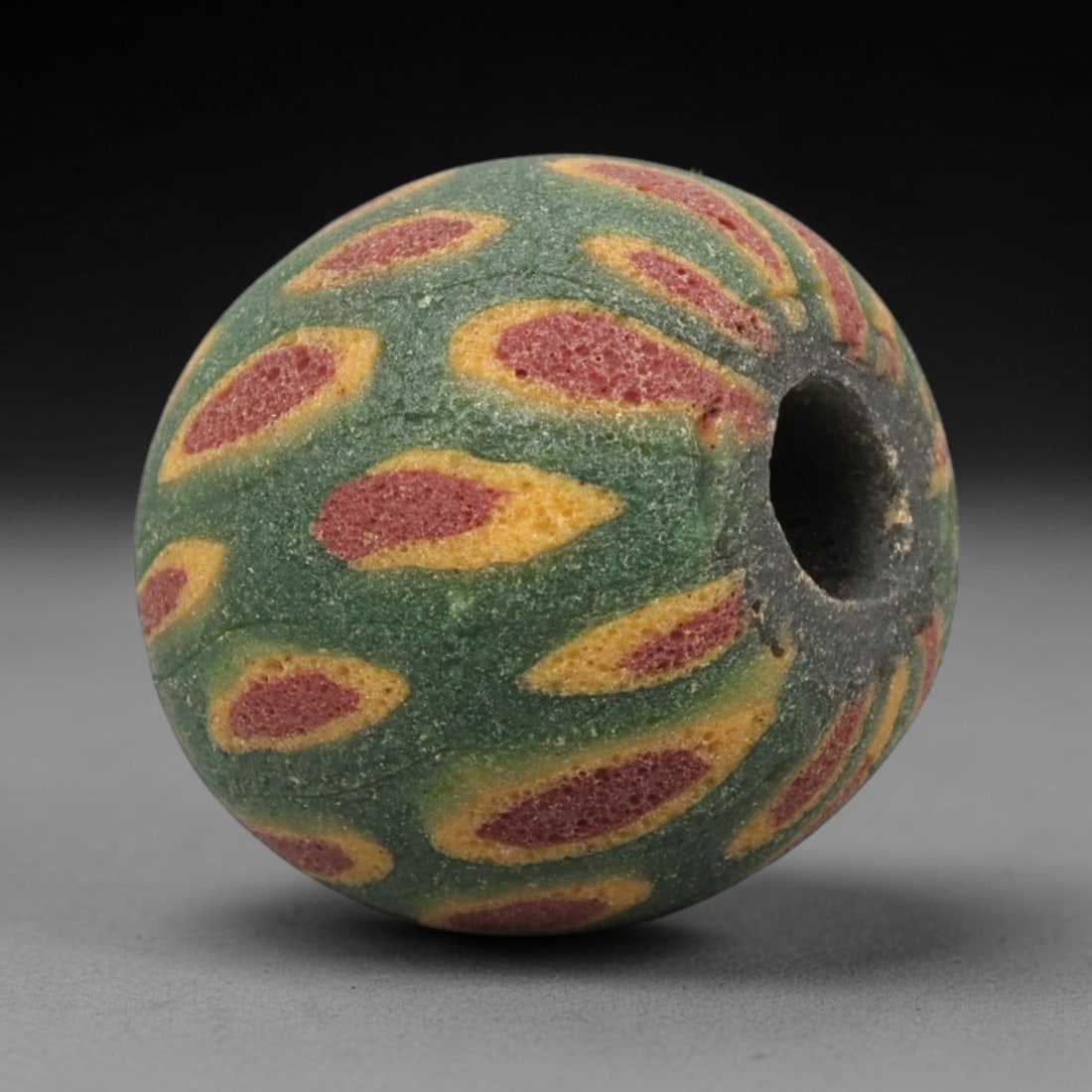 Ancient Mosaic Glass Bead - Roman Floral Pattern Amulet, 69WD: This spherical bead is crafted from core-formed mosaic glass, featuring a deep green base decorated with repeating teardrop-shaped floral motifs. Each motif consists of a central red glass inlay outli