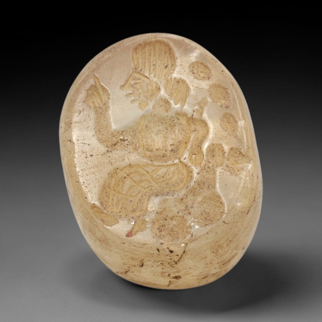 Ancient Rock Crystal Stamp Seal - Near Eastern Anthropomorphic Figure, 63ZR: Carved from translucent pale rock crystal, this oval intaglio stamp seal depicts a seated anthropomorphic figure in raised relief, with stylized linear details defining the figure's headdress, torso,