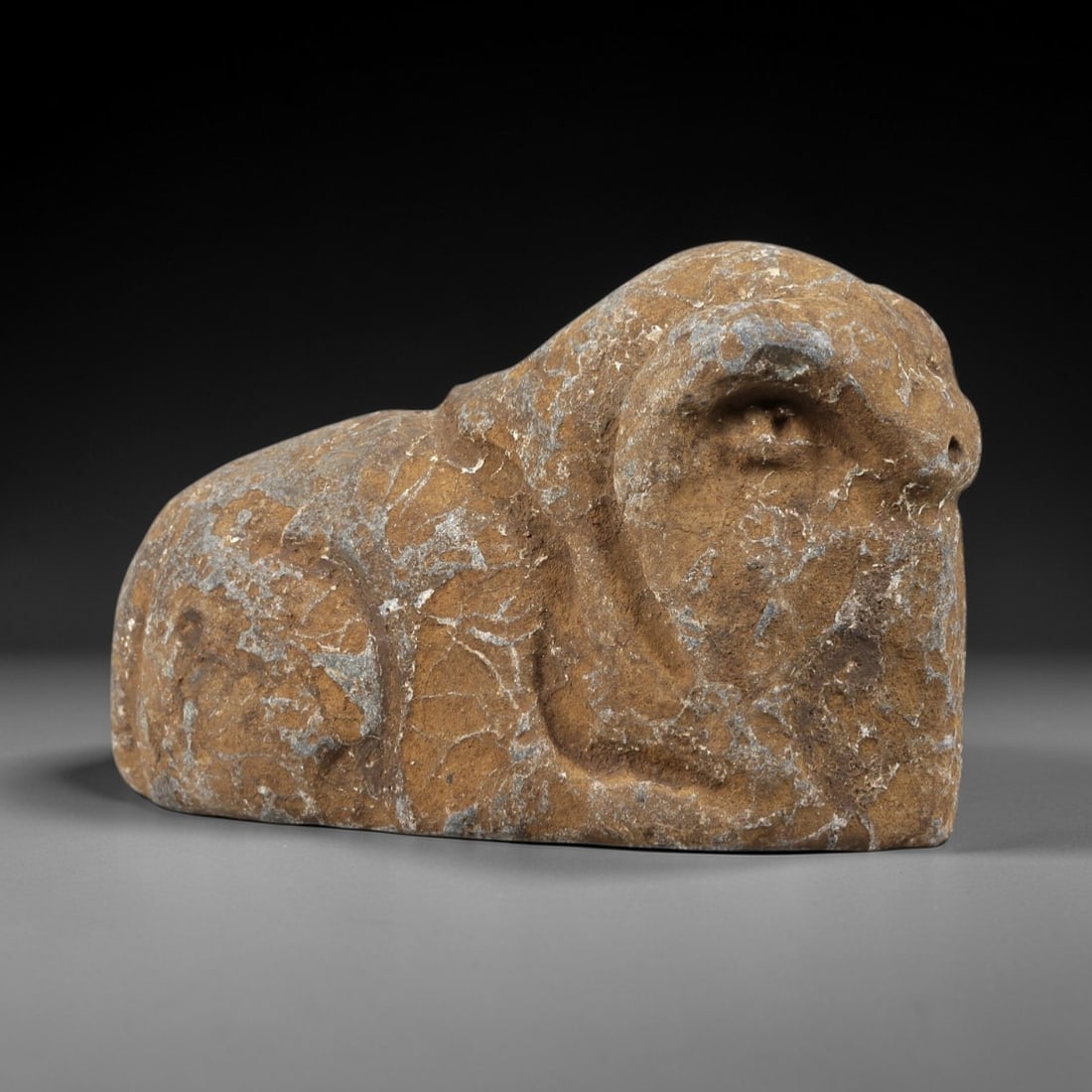Ancient Stone Ram Effigy - Near Eastern Zoomorphic Figurine, 97FX: Carved from a weathered, veined stone, this effigy depicts a recumbent ram in a compact, stylized form. Relief details define the curled horns, snout, and rounded body, emphasizing the animal's robust