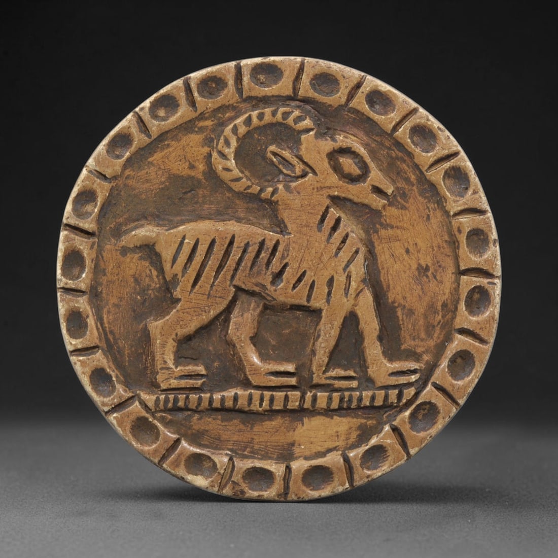 Ancient Steatite Plaque - Near Eastern Ram Relief, 34ZY: Carved from steatite, this circular plaque features a relief depiction of a stylized ram, rendered in the schematic artistic tradition of the Ancient Near East. The animal is shown walking in profile,