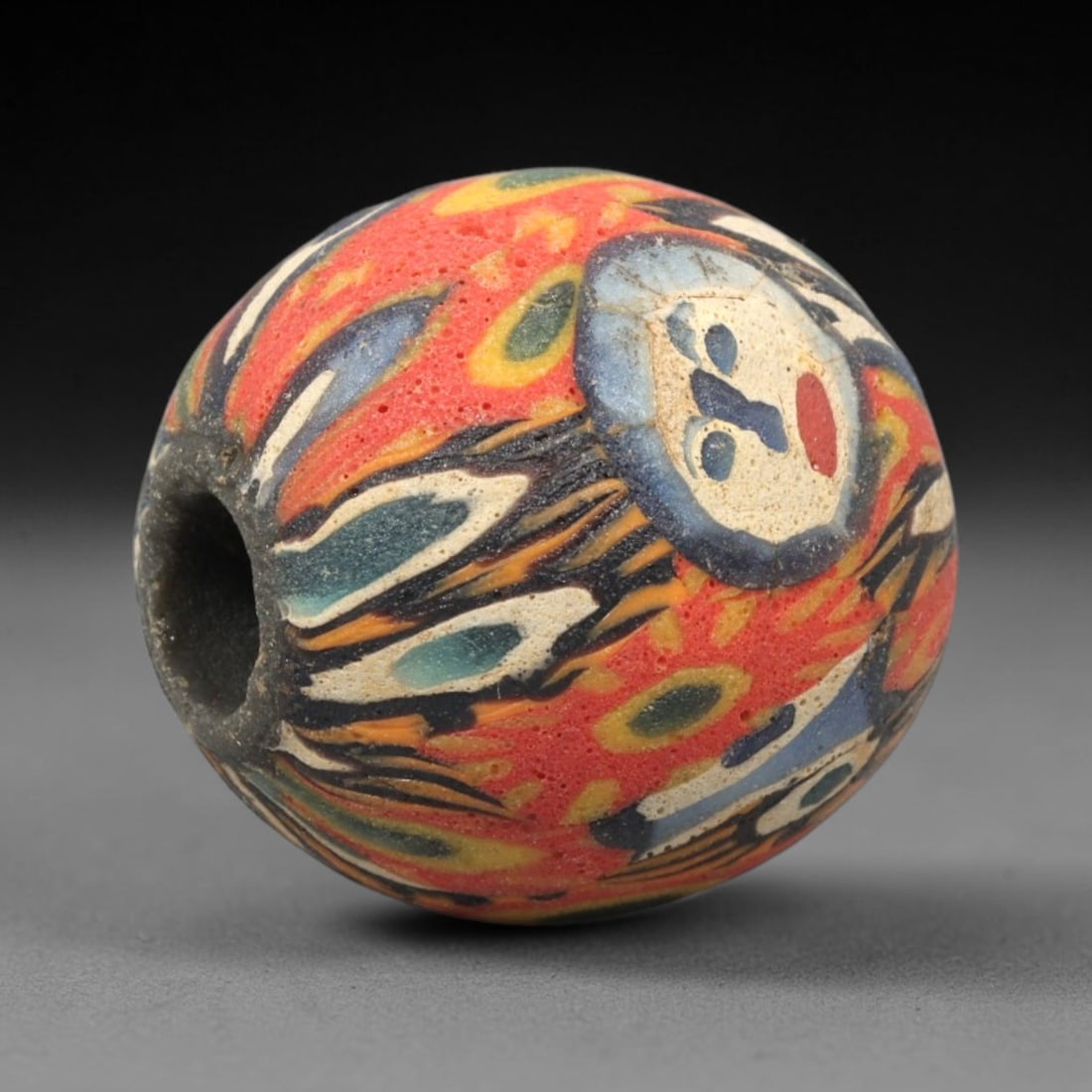 Ancient Mosaic Glass Bead - Roman Face Pattern Amulet, 54WD: This spherical bead is crafted from core-formed mosaic glass, featuring a vibrant red base decorated with intricate face motifs and swirling foliate elements. Each face motif consists of a cream-color