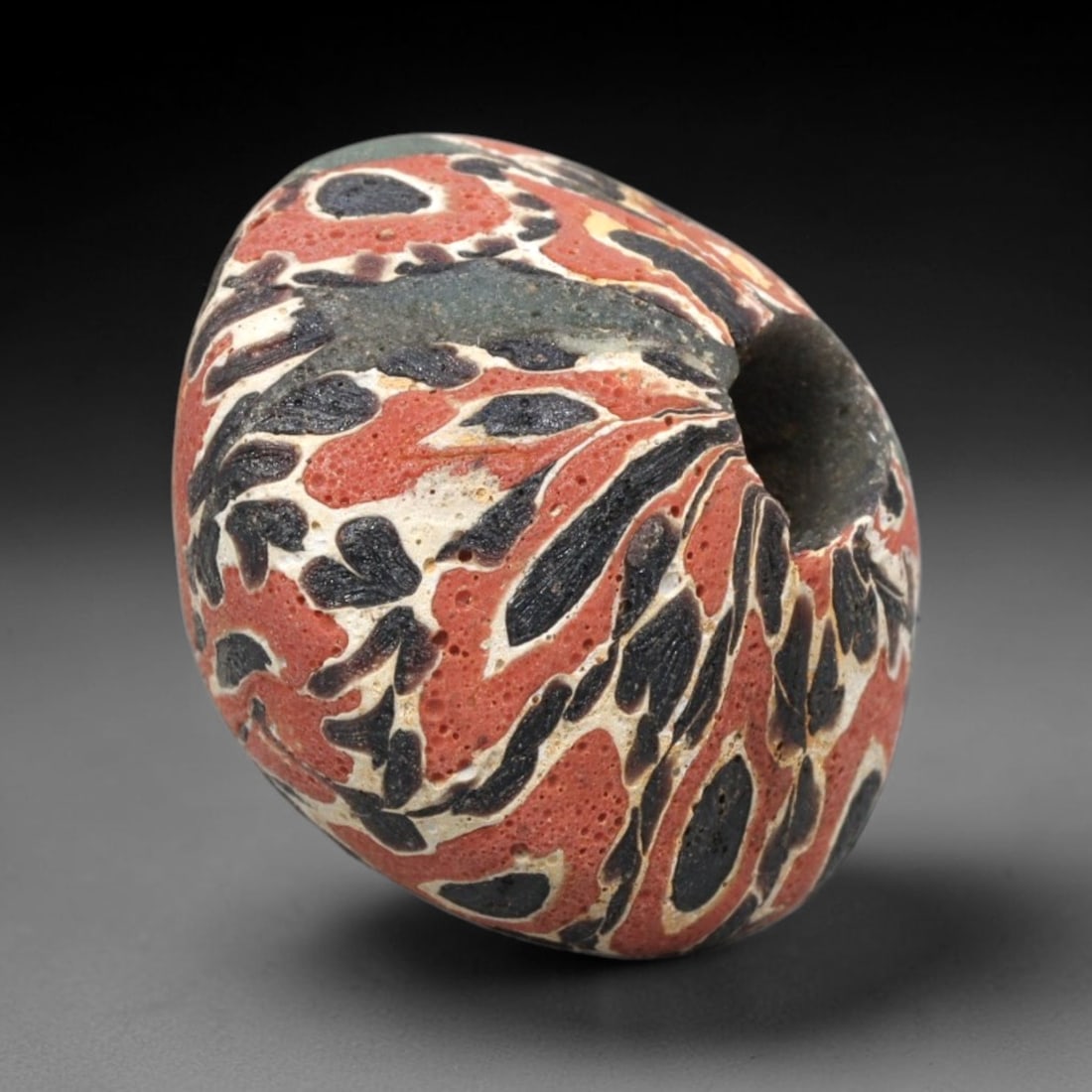 Ancient Mosaic Glass Bead - Roman Floral Pattern Amulet, 64WD: This barrel-shaped bead is crafted from mosaic glass, featuring a vibrant red base decorated with intricate layered glass canes in black, white, and pale yellow. The decorative pattern consists of swi