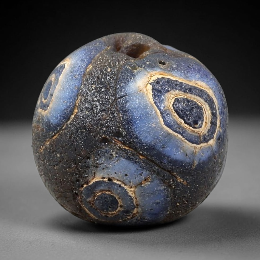 Ancient Glass Bead - Roman Mosaic Eye Bead Pendant, 20BG: A spherical ancient glass bead crafted in the mosaic glass technique, with a deep blue glass base adorned with concentric "eye" motifs, each formed by layered blue and cream-colored glass canes. The s