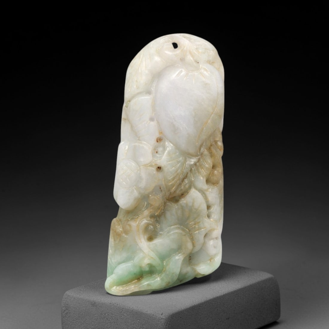 A Chinese Jadeite Jade Floral Pendant, 20th Century, 99FR: Carved from variegated jadeite jade with natural white, celadon green, and brownish tonal variations, the pendant has an elongated, irregular freeform shape with a drilled suspension hole at the apex.
