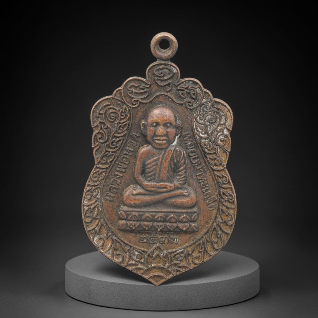 A Thai Copper Alloy Buddhist Amulet, Late 20th Century, 95FP: A shield-shaped copper alloy amulet, measuring 4.5 x 3.8 cm. The obverse depicts a seated Buddhist monk in meditative mudra, resting on a lotus pedestal, with Thai script inscriptions encircling the f