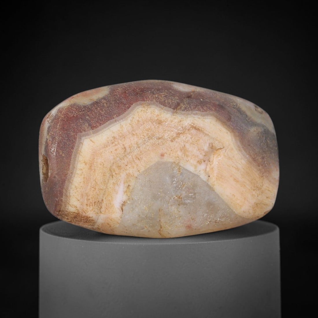 Ancient Agate Bead - Bactrian Artifact, 24BD: This artifact is carved from layered agate, formed into a broad, flattened ovoid shape with distinct concentric banding in earthy red and cream hues. The bead features a central perforation for suspen