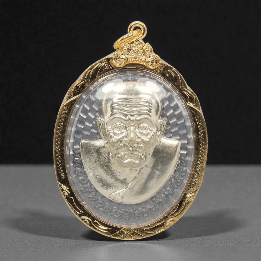 A Thai Silver-Toned Metal Monk Portrait Amulet with Gilt-Metal Frame, 21st Century, 63FP: The amulet is of oval form, featuring a high-relief silver-toned bust portrait of a Buddhist monk on the obverse, set against a radiating linear ground with incised Thai script arranged in a circular 