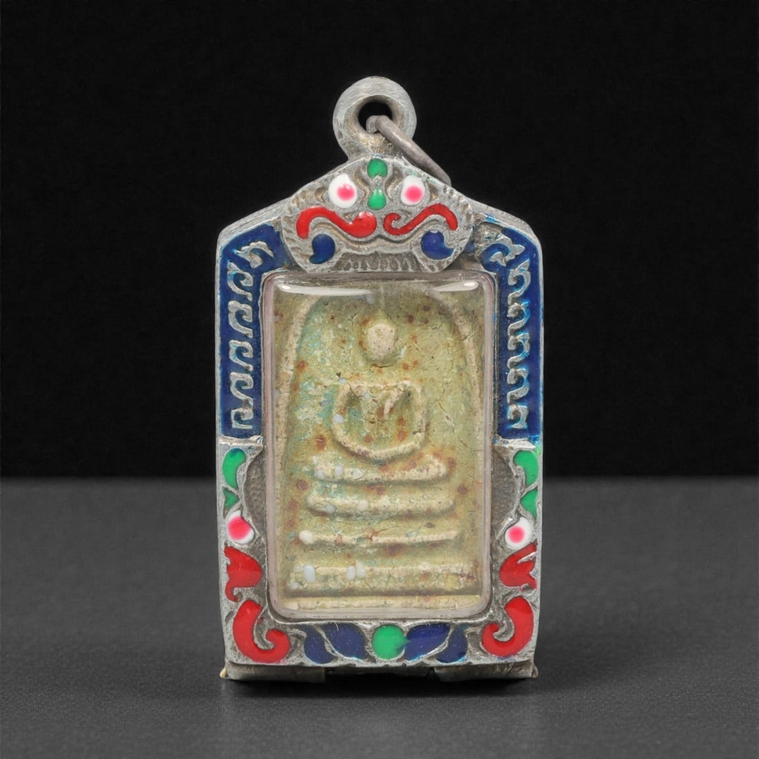 A Thai Clay Buddha Amulet in Silver Enamel Case, 20th Century, 20FP: Molded from light greenish-tan clay with embedded white inclusions and surface patination, the amulet depicts a seated Buddha in meditative posture atop a multi-tiered base. The reverse is plain, with