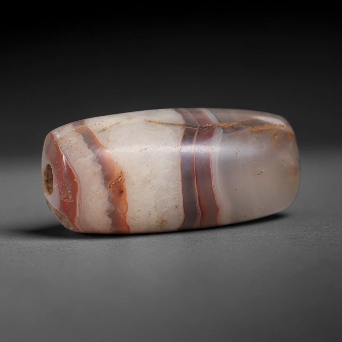 Ancient Agate Bead - Bactrian Amulet, 11BD: This artifact is crafted from banded agate, showcasing bold alternating stripes of deep red, creamy white, and translucent grey, shaped into a smooth elongated barrel form through skilled ancient lapi