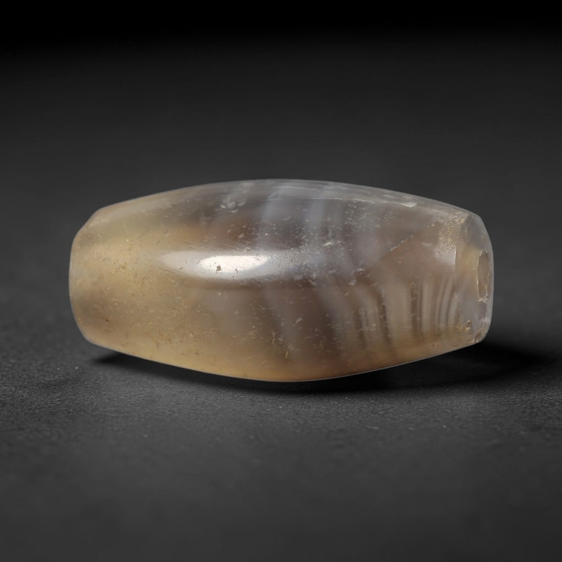 Ancient Agate Bead - Near Eastern Amulet, 14BD: This artifact is crafted from translucent banded agate, exhibiting subtle layered striations in pale grey, warm amber, and soft brown tones, shaped into a smooth faceted barrel form through skilled an