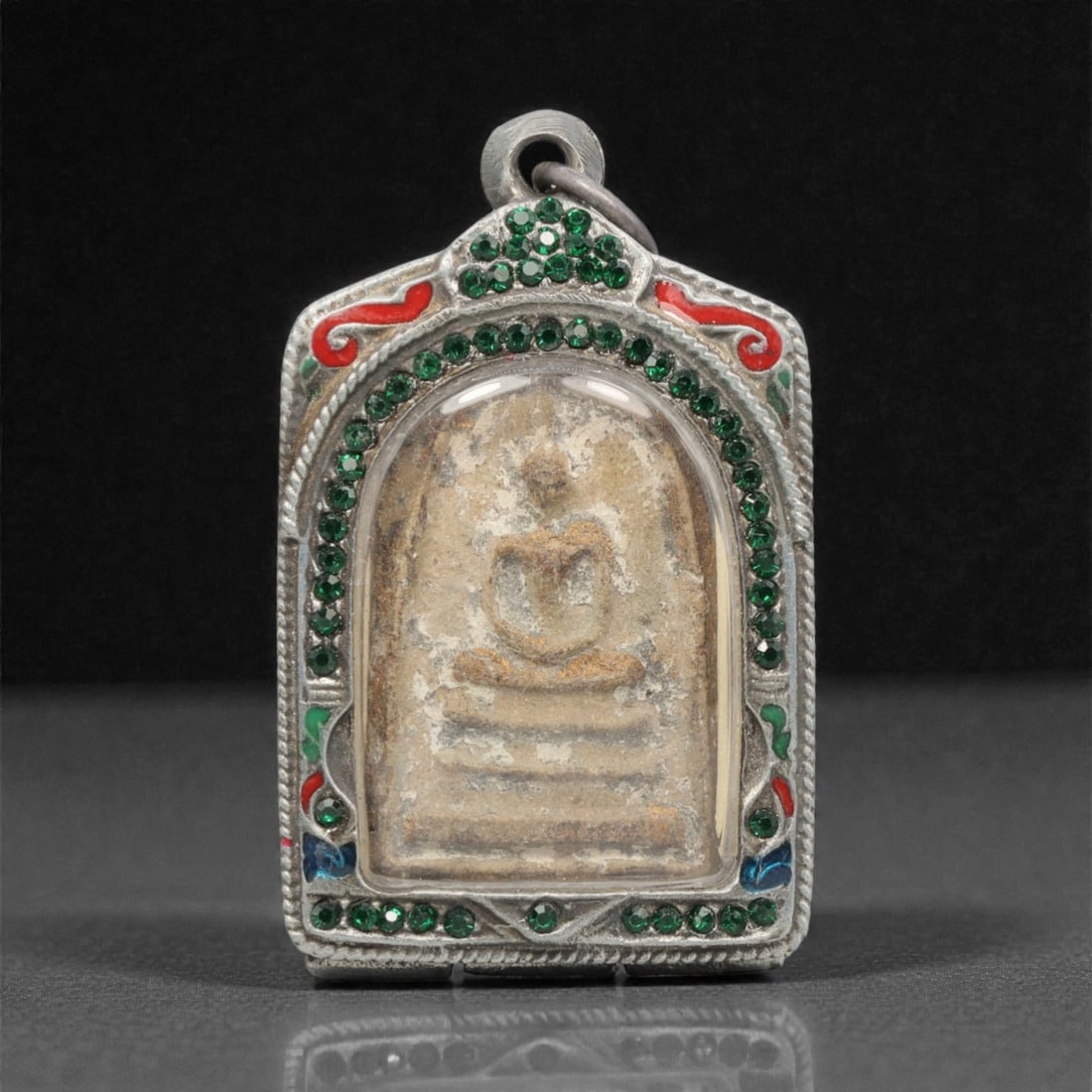 A Thai Clay Buddha Amulet in Silver Enamel and Gem-Set Case, 20th Century, 99FN: Molded from light tan clay with extensive surface patination and wear, the amulet depicts a seated Buddha in meditative posture atop a multi-tiered base. The reverse is heavily patinated, with indisti