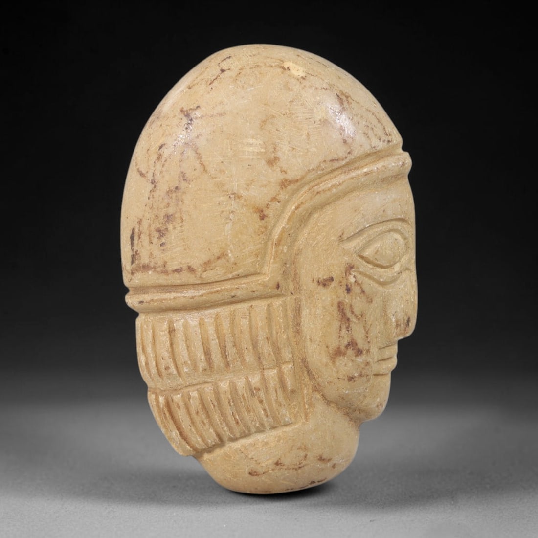 Ancient Stone Head Amulet - Near Eastern Bust Plaque, 32FY: Carved from a pale beige limestone with subtle brown veining, this amulet takes the form of a three-dimensional human head bust in high relief, crafted in the iconographic tradition of ancient Near Ea