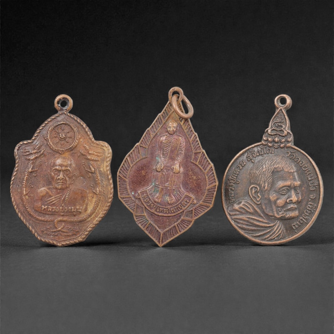 Three Thai Copper Alloy Devotional Amulets, 20th Century, 74FN: Cast from copper alloy with a rich brown patina, the group comprises three distinct devotional amulets, each with a suspension loop at the apex for wear. The first amulet is shield-shaped, depicting a