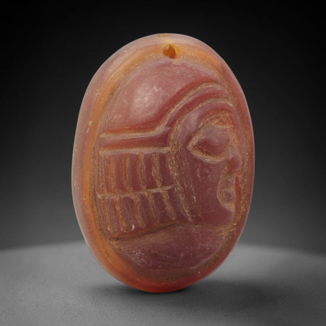 Ancient Carnelian Amulet - Near Eastern Bust Pendant, 33ZX: Carved from carnelian, the amulet is an oval disc with a drilled suspension hole at the top, crafted in the ancient Near Eastern style. The front surface bears a raised relief portrait of a human bust