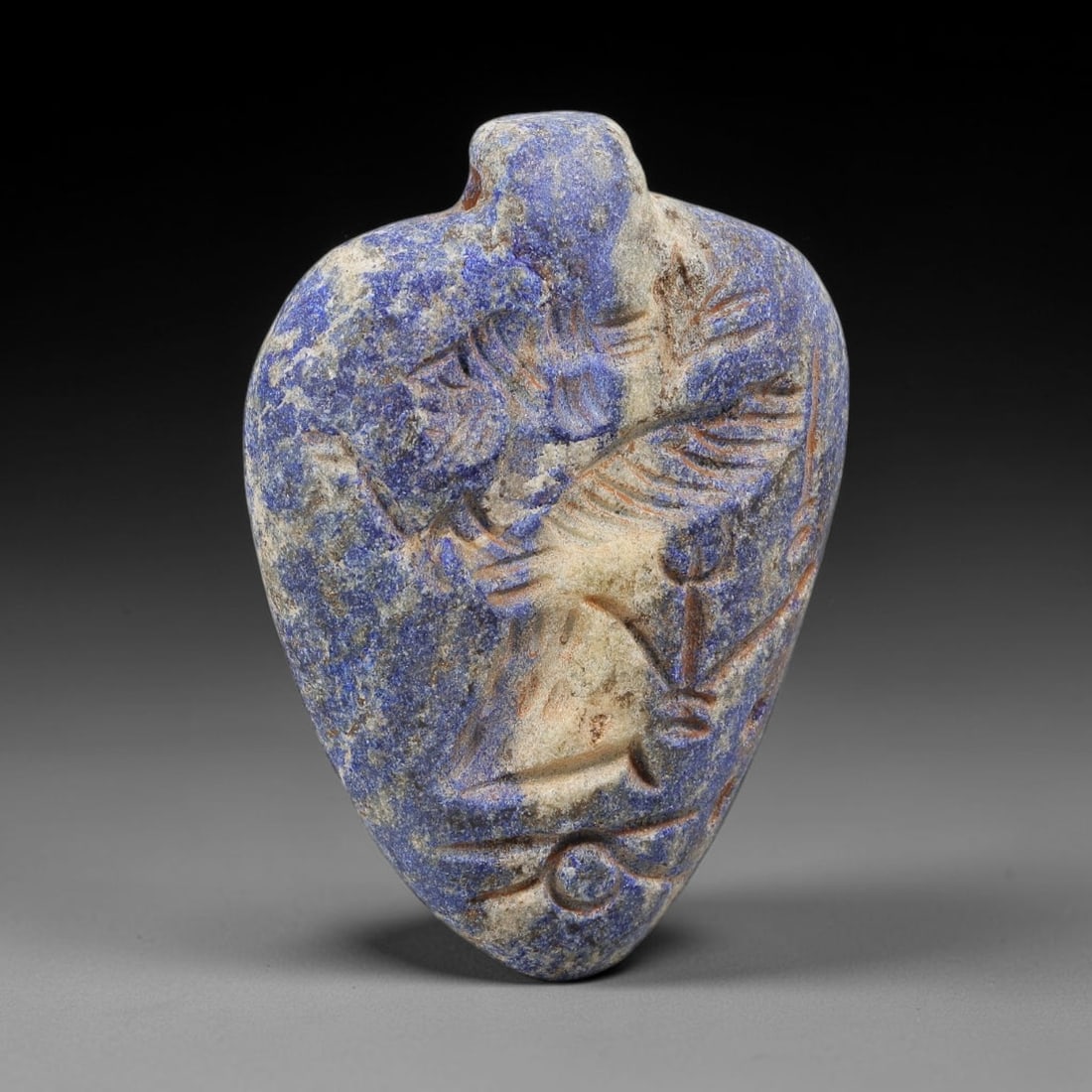 Ancient Lapis Lazuli Amulet - Near Eastern Anthropomorphic Figurine, 40BA: Carved from lapis lazuli, the amulet takes a heart-shaped form with a suspension loop at the top, designed for wear as a protective talisman. The front face is incised with a stylized anthropomorphic 