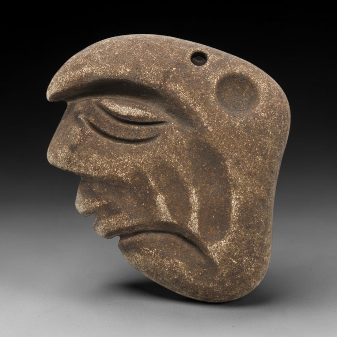 Ancient Stone Anthropomorphic Face - Near Eastern Amulet, 90ZV: This artifact is a carved stone anthropomorphic face amulet, rendered in a granular, earth-toned stone consistent with ancient Near Eastern lithic traditions. The relief carving depicts a stylized pro