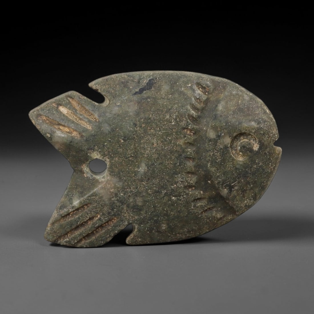 Ancient Stone Fish Amulet - Egyptian Ritual Figurine, 53AE: This amulet is carved from a single piece of mottled green-grey stone, exhibiting a fine-grained texture with subtle lighter mineral inclusions and natural veining across its surface. The form is styl