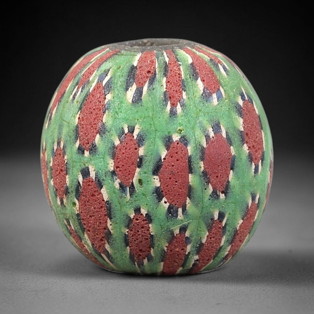 Ancient Glass Bead - Roman Millefiori Spherical Pendant, 37BG: A spherical ancient glass bead crafted in the millefiori mosaic technique, with a vibrant green glass base overlaid with intricate fused cane patterns. The surface is decorated with repeating teardrop
