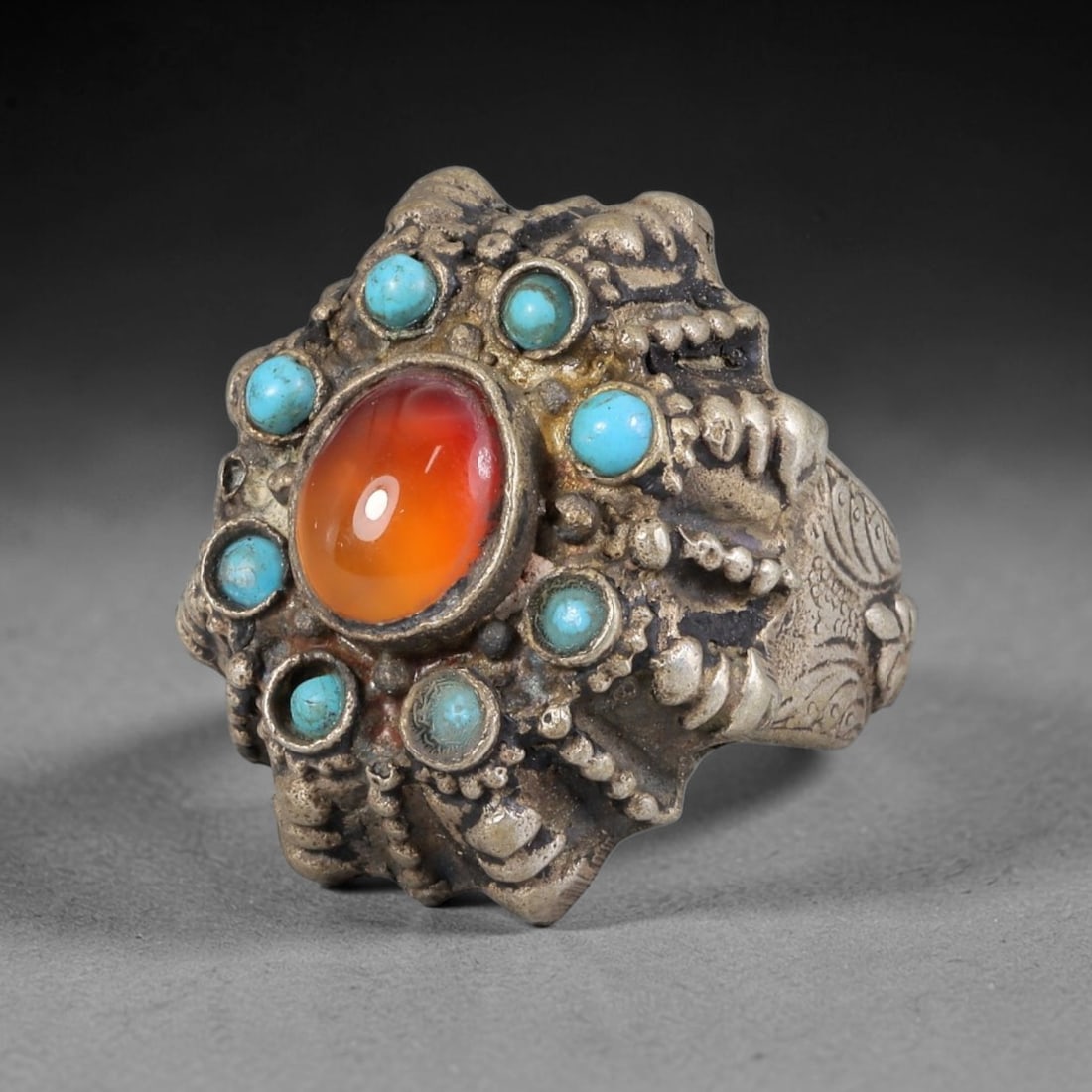 Antique Silvered Ring - Eurasian Steppe Carnelian Turquoise Cluster, 31BK: Silvered metal ring centered with a large oval carnelian cabochon displaying rich orange-red hues, surrounded by a halo of round turquoise cabochons. The ring features elaborate repousse and pseudo-fi