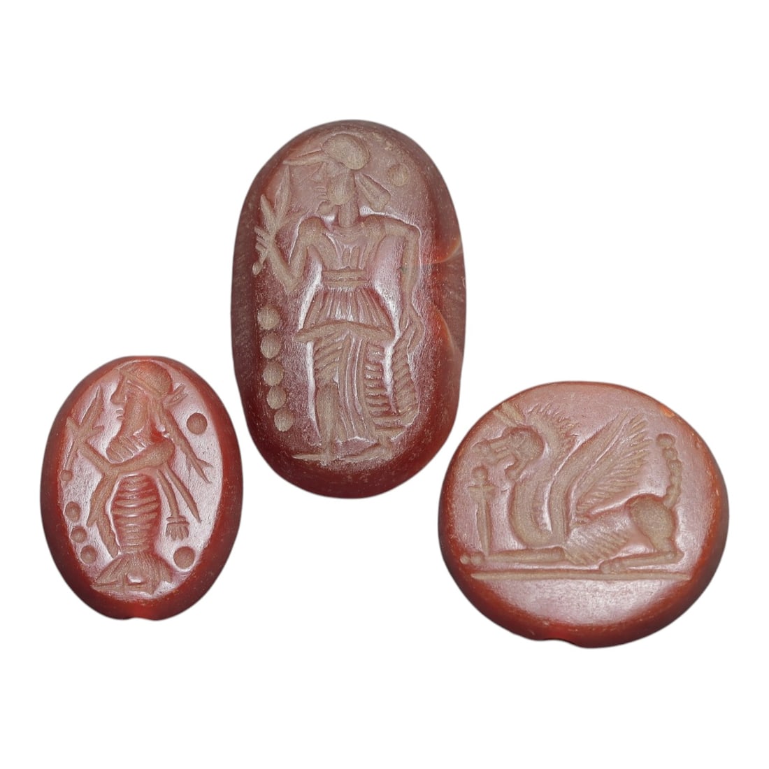 Lot of 3 Ancient Carnelian Intaglio Seals, Greco-Persian, Circa 5th-3rd Century BCE, 100AD: Comprising three carnelian intaglio seals of varying form and iconography. The largest oval seal depicts a standing figure in profile, wearing a headdress and layered skirt, holding a palm branch and 
