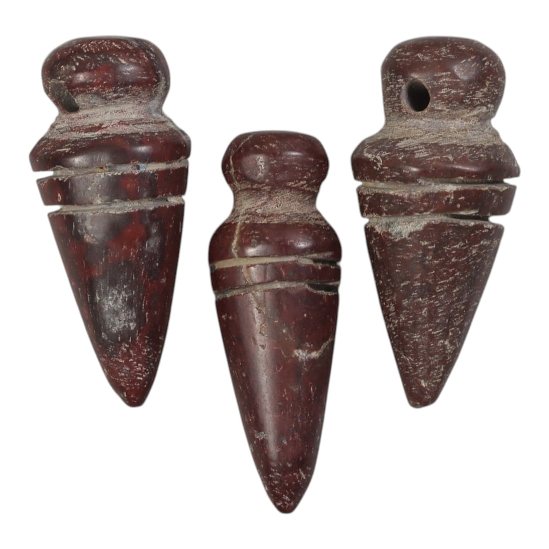 Lot of 3 Ancient Coptic Egyptian Red Stone Amulets, Circa Roman to Coptic Era, 40AE: The group comprises three carved red stone amulets of stylized conical form. Each features a rounded suspension lug at the top, with incised bands below and a tapering pointed lower section. The stone