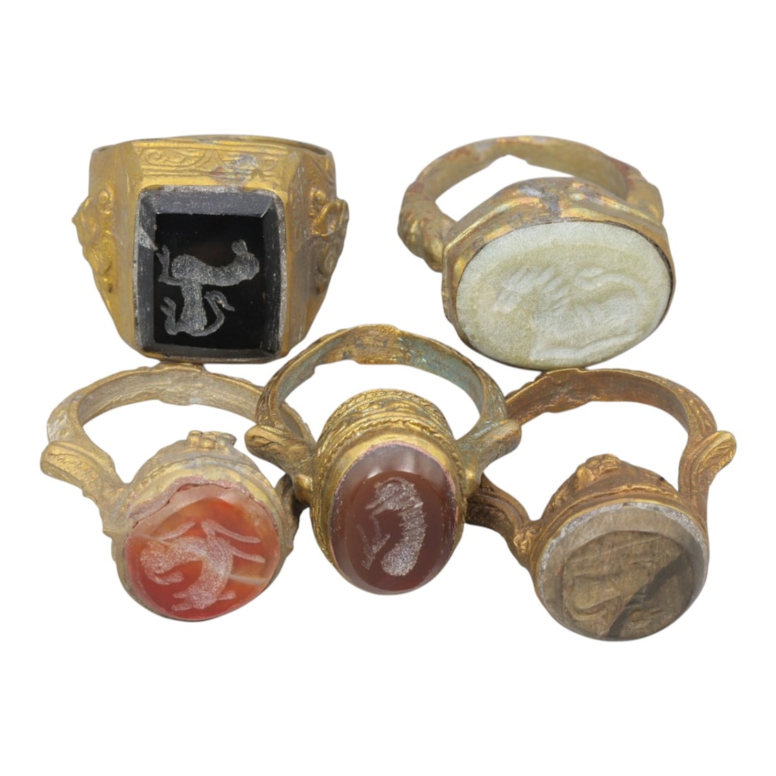 Lot of 5 Antique Qajar Period Gilt Intaglio Seal Rings, 4AD: A collection of five antique gilt-metal rings from the Qajar period (19th century), each featuring a carved intaglio seal stone. The set includes varied gemstone materials and motifs: one black onyx w