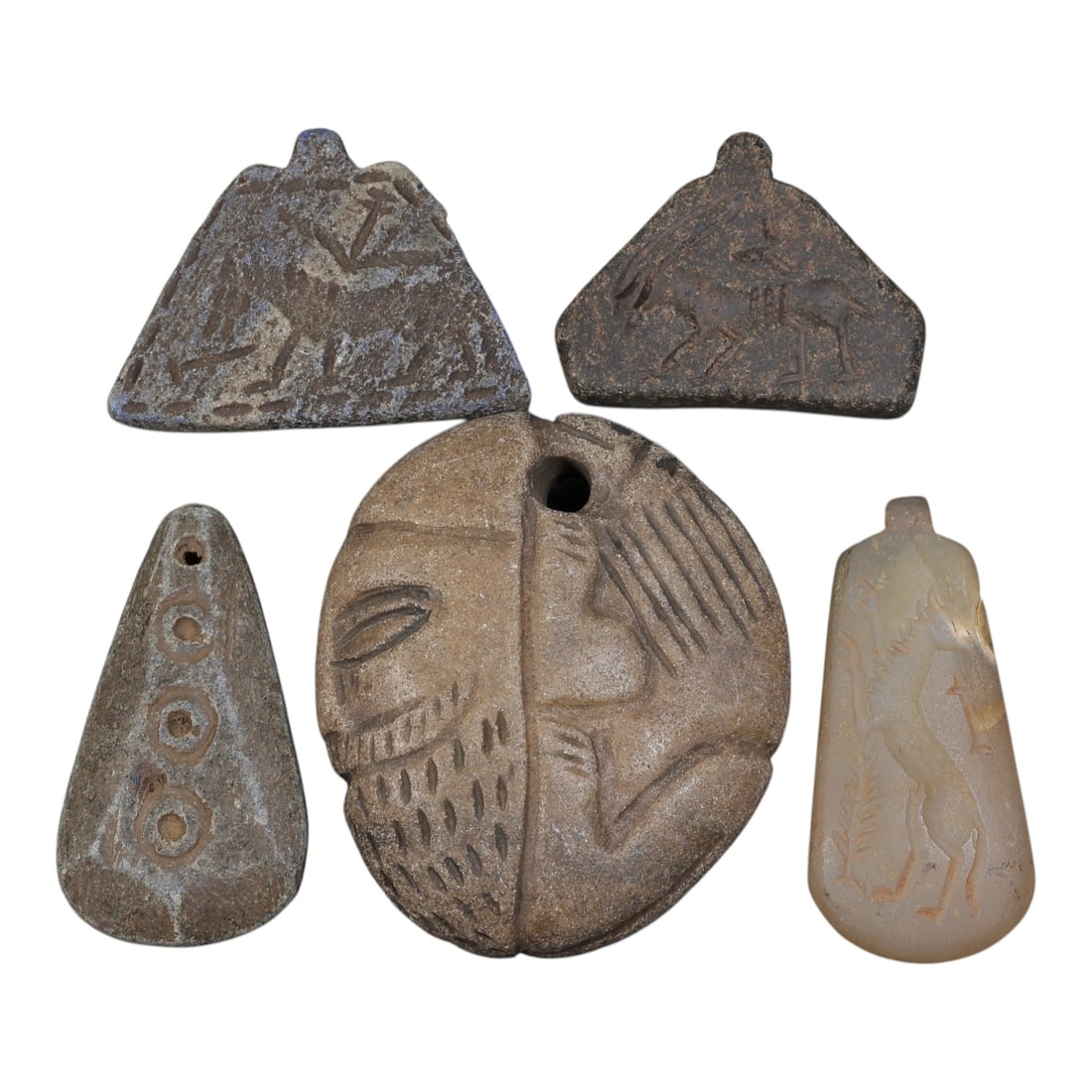 Lot of 5 Ancient Near Eastern Stone Pendants and Intaglios, Circa 1st Millennium BCE, 93AD: Comprising five stone pendants and intaglios of varying form, material, and iconography. Two triangular dark stone pendants with zoomorphic incised designs depicting quadrupeds, likely ibex or gazelle