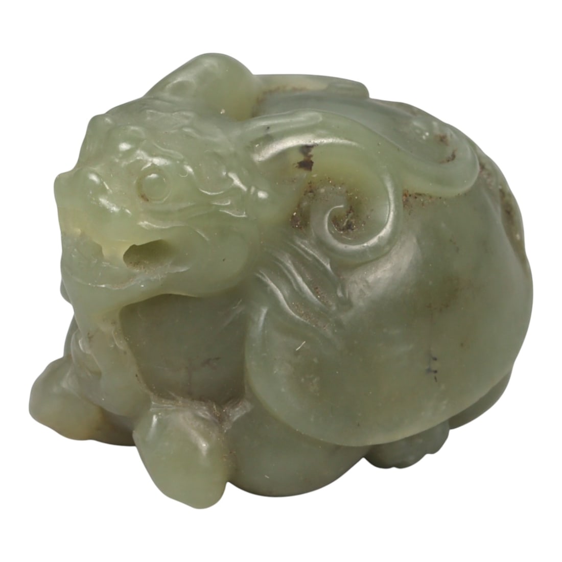 A Chinese Nephrite Jade Pixiu Figure, 20th Century, 3YA: Carved from pale green nephrite jade with soft translucency and scattered natural inclusions, the figure represents a pixiu shown in a compact, crouching posture. The head is strongly modeled with a p