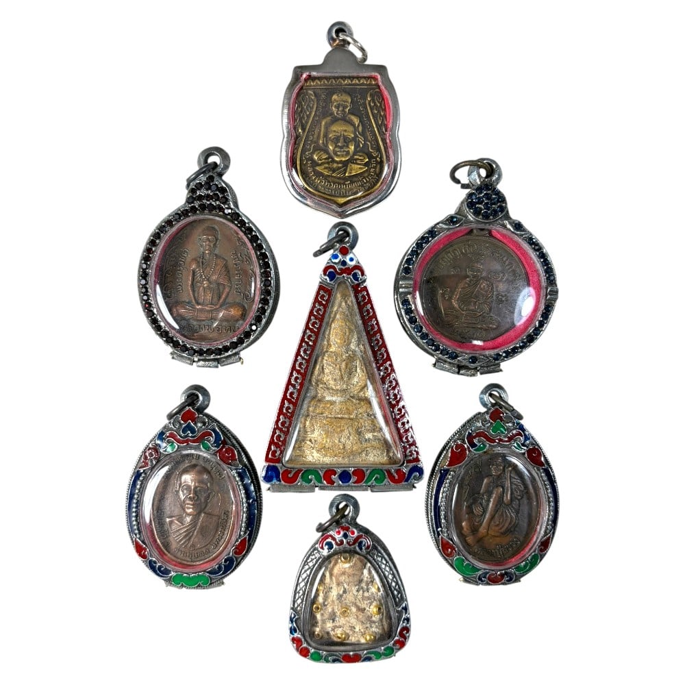 A Lot of 7 Vintage Thai Buddhist Amulets with Enamel and Jewel Accented Cases, 25AA: Comprising seven pendants ranging from 4 to 7 cm in height. The collection features a mix of aged monastic portrait medallions and sacred Buddha images, encased in ornate silver-toned metal cases. The