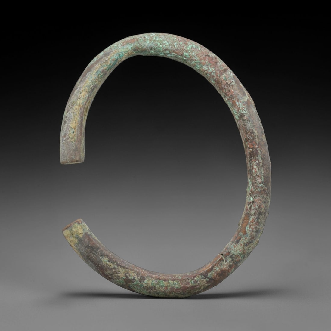 A Roman Bronze Bracelet, 1st-3rd Century CE, 96FT: Cast from bronze with a thick, rounded section, the bracelet is formed as an open C-shape with flat, slightly tapered terminals. The surface is covered in a patchy green patina with extensive reddish-
