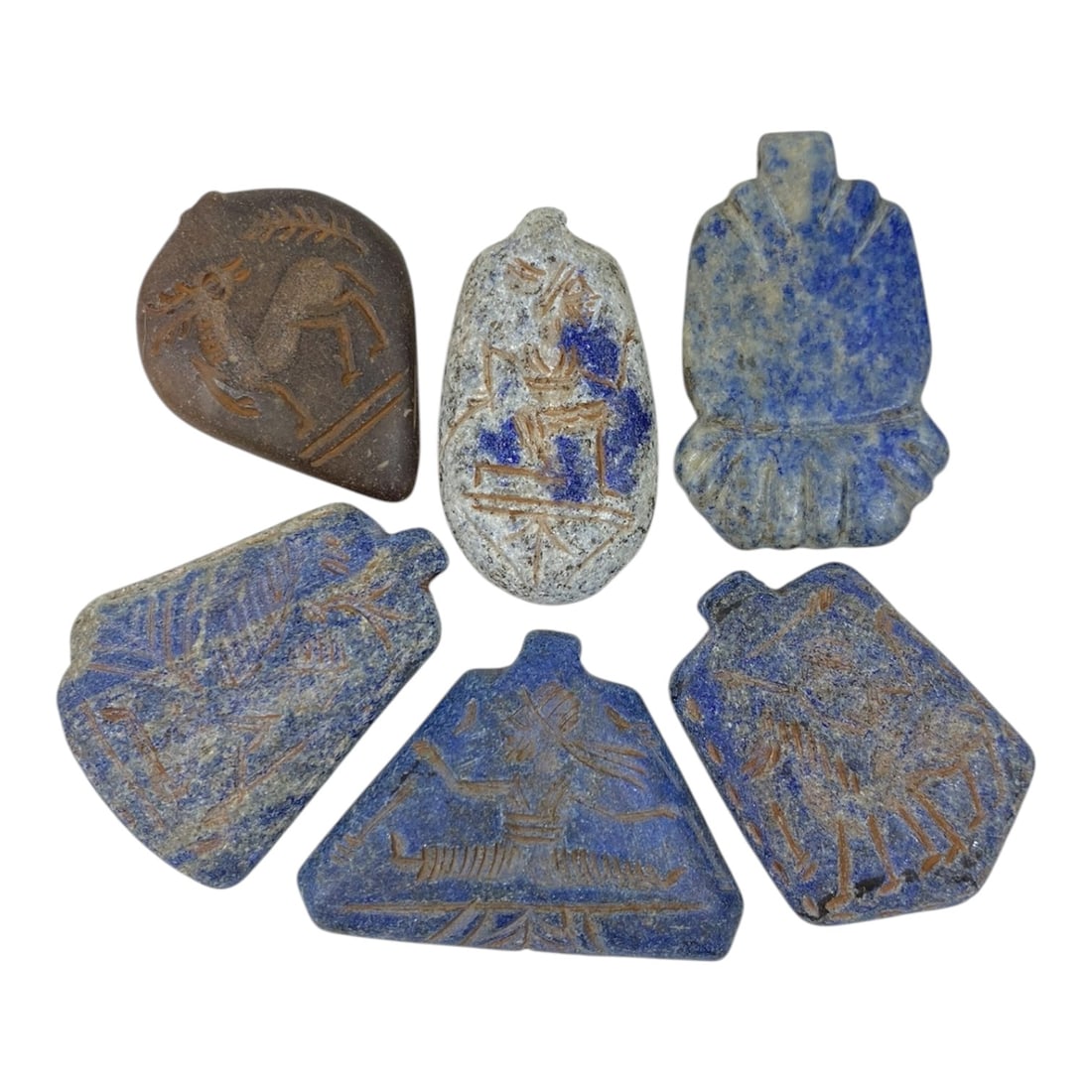 A Lot of 6 Ancient Near Eastern Lapis Lazuli Amulets, 10AC: The group comprises six amulets, five carved from lapis lazuli and one from a brown stone, exhibiting varied tones and natural mineral inclusions, alongside surface patina and wear consistent with ant