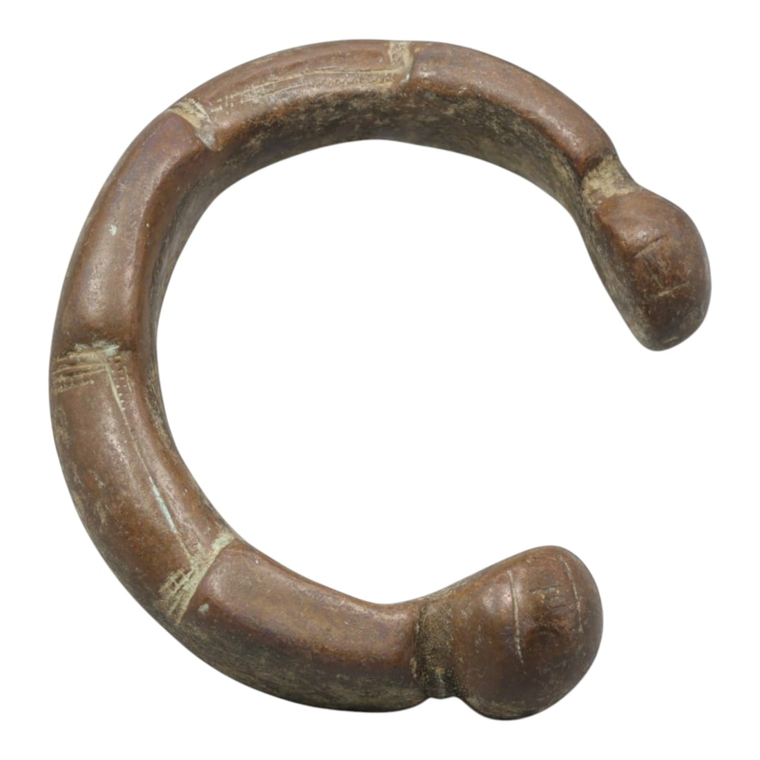 An African Bronze Manila Bracelet, 19th Century, 99FT: Cast from bronze with a thick, rounded section, the bracelet is formed as an open C-shape with prominent, bulbous terminals. The band is segmented by shallow, incised horizontal lines, creating a seri