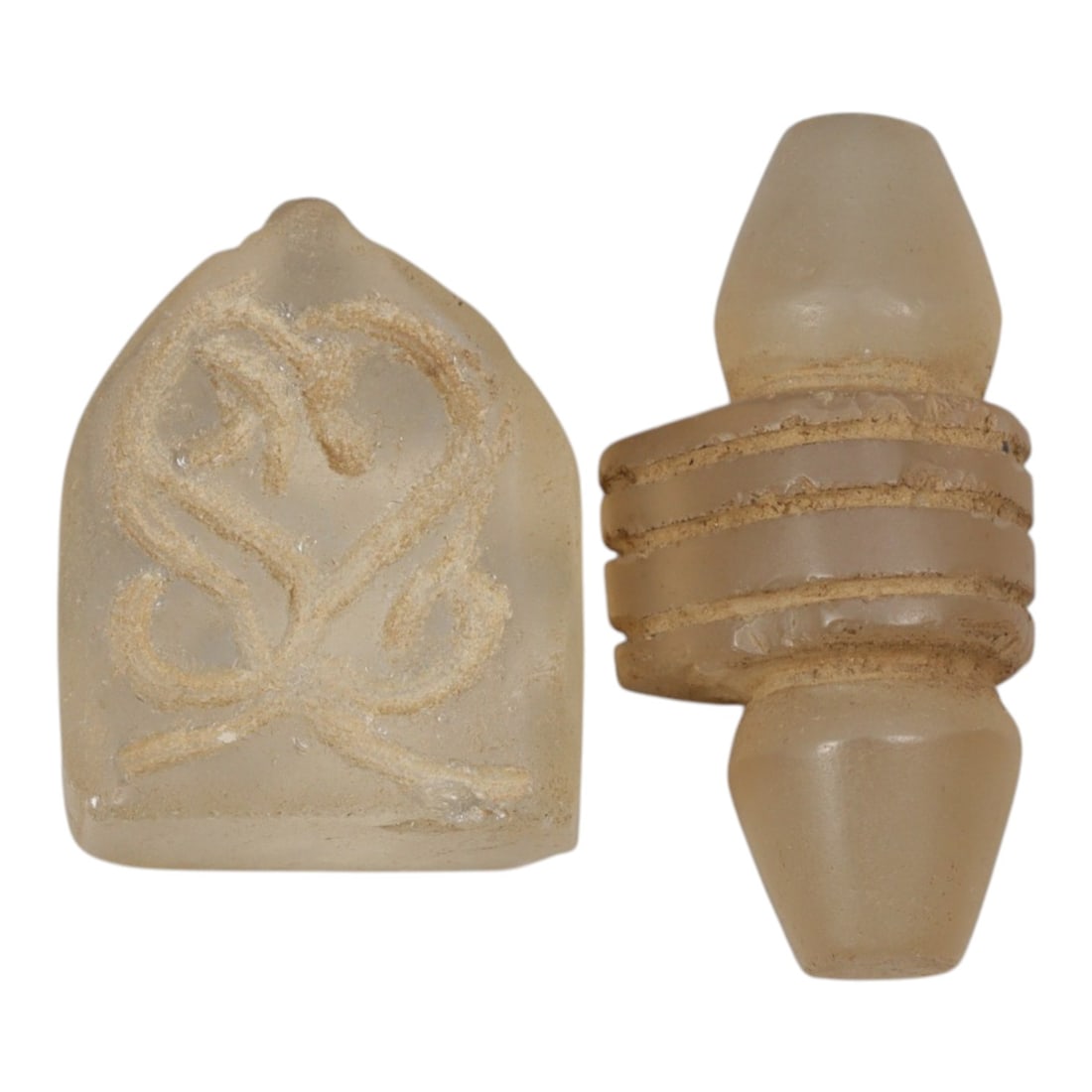 Lot of 2 Ancient Medieval Egyptian Glass Amulets, c. Late Antique to Islamic Period, 19WL: A pair of translucent pale glass amulets from Ancient Medieval Egypt. The first is a shield-shaped pendant with incised scrollwork motifs, while the second is a stylized columnar bead with raised band