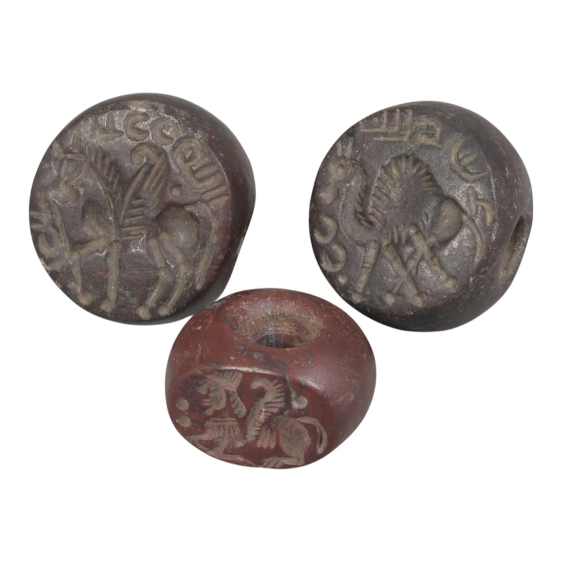 Lot of 3 Ancient Sasanian Stone Stamp Seals, Circa 3rd-7th Century CE, 66WP: A collection of three Sasanian period stamp seals, carved from reddish-brown stone, each featuring a central drilled hole for suspension. The seals display characteristic Sasanian iconography: includi