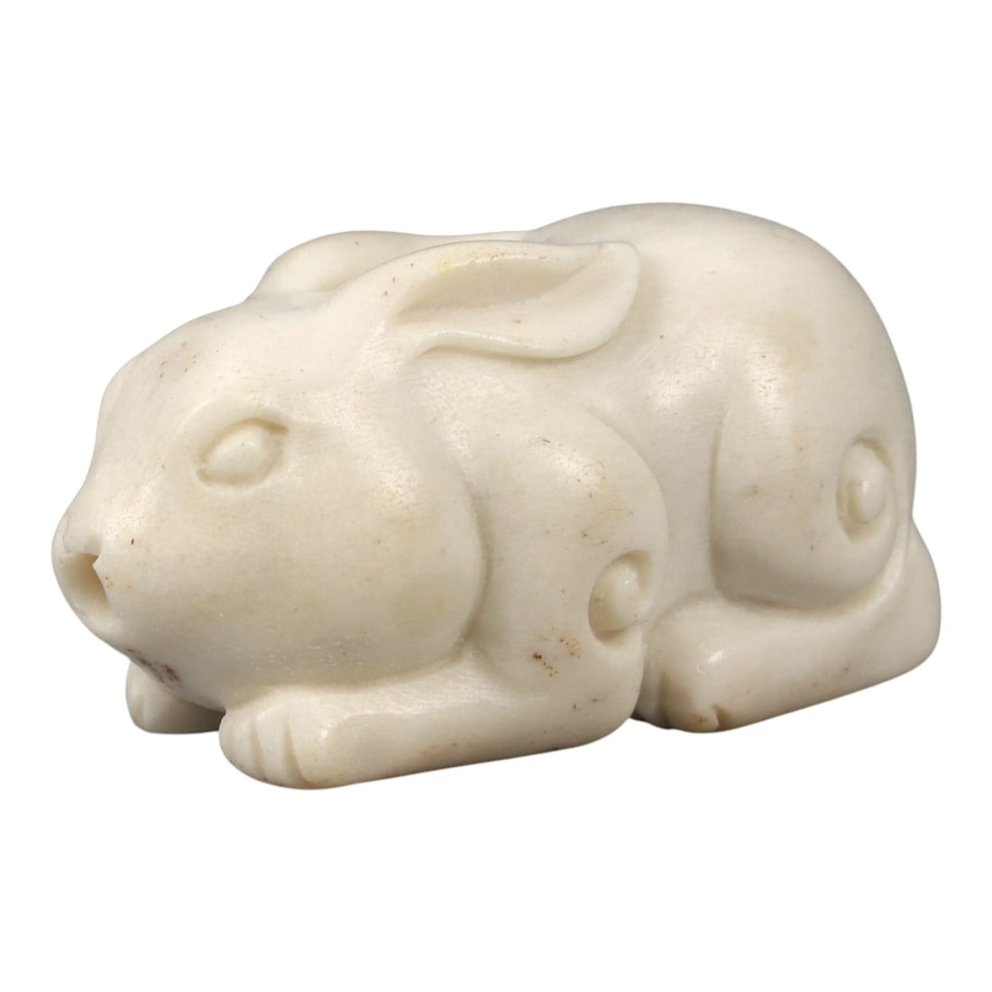 An East Asian Carved Rabbit Bead, 20th Century, 5FD: Carved from a pale, opaque material of off-white tone, the bead is modeled in the form of a recumbent rabbit with legs folded beneath the body. The head is rendered with a rounded snout, shallowly def