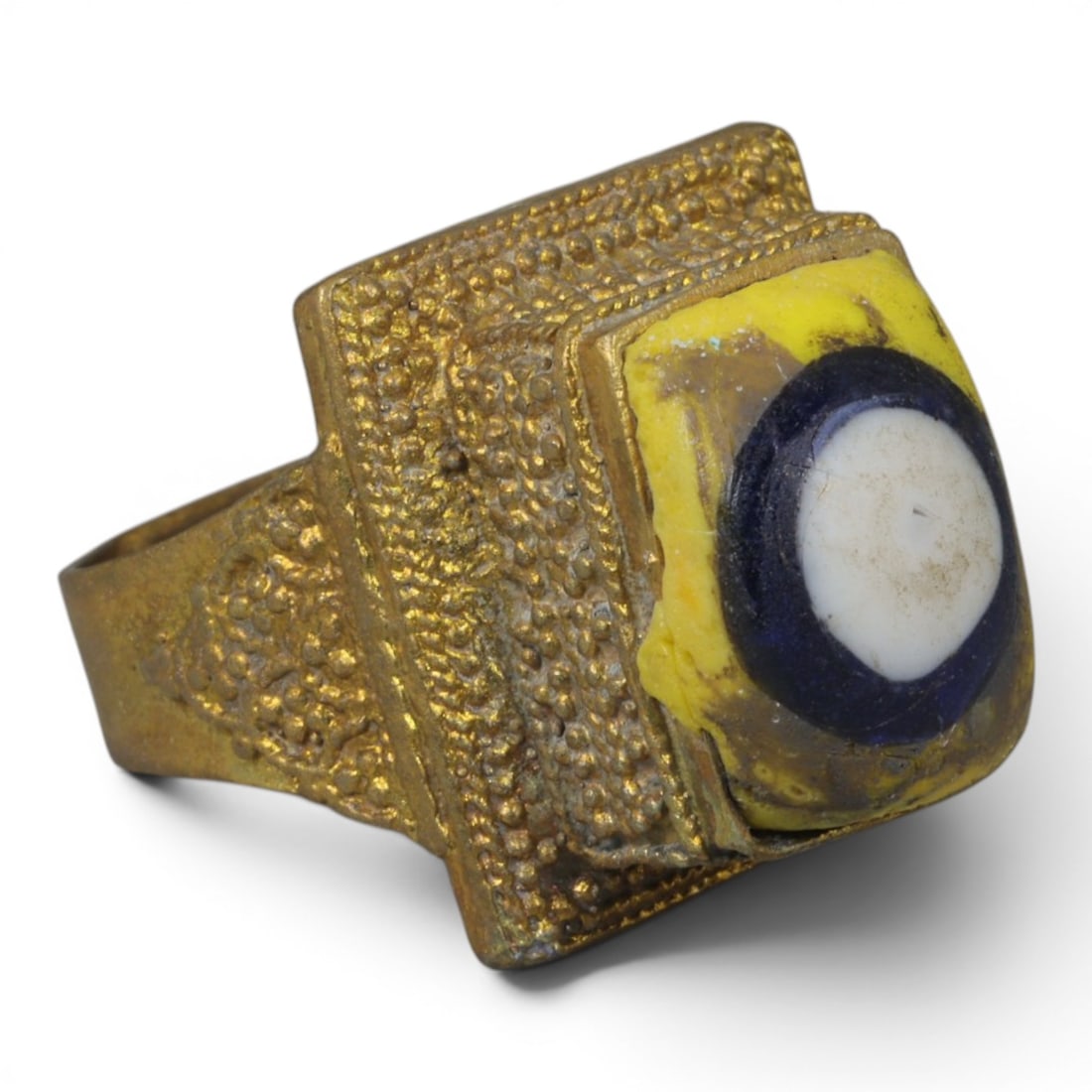 Antique Qajar Dynasty Gilt Evil Eye Ring, US Size 8, 53AJ: A gilt-metal ring of the Qajar period, US ring size 8, featuring a large square evil-eye glass inlay with a vibrant yellow, blue, and white concentric design. The bezel and band are adorned with intri