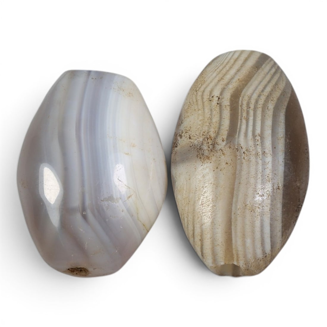 A Pair of Antique Central Asian / Himalayan Banded Agate Trade Beads, 15th-19th Century, 85AJ: The pair comprises two large, ovoid banded agate beads. The left bead displays soft, translucent grey and white banding with a smooth, polished surface, while the right bead features distinct, paralle