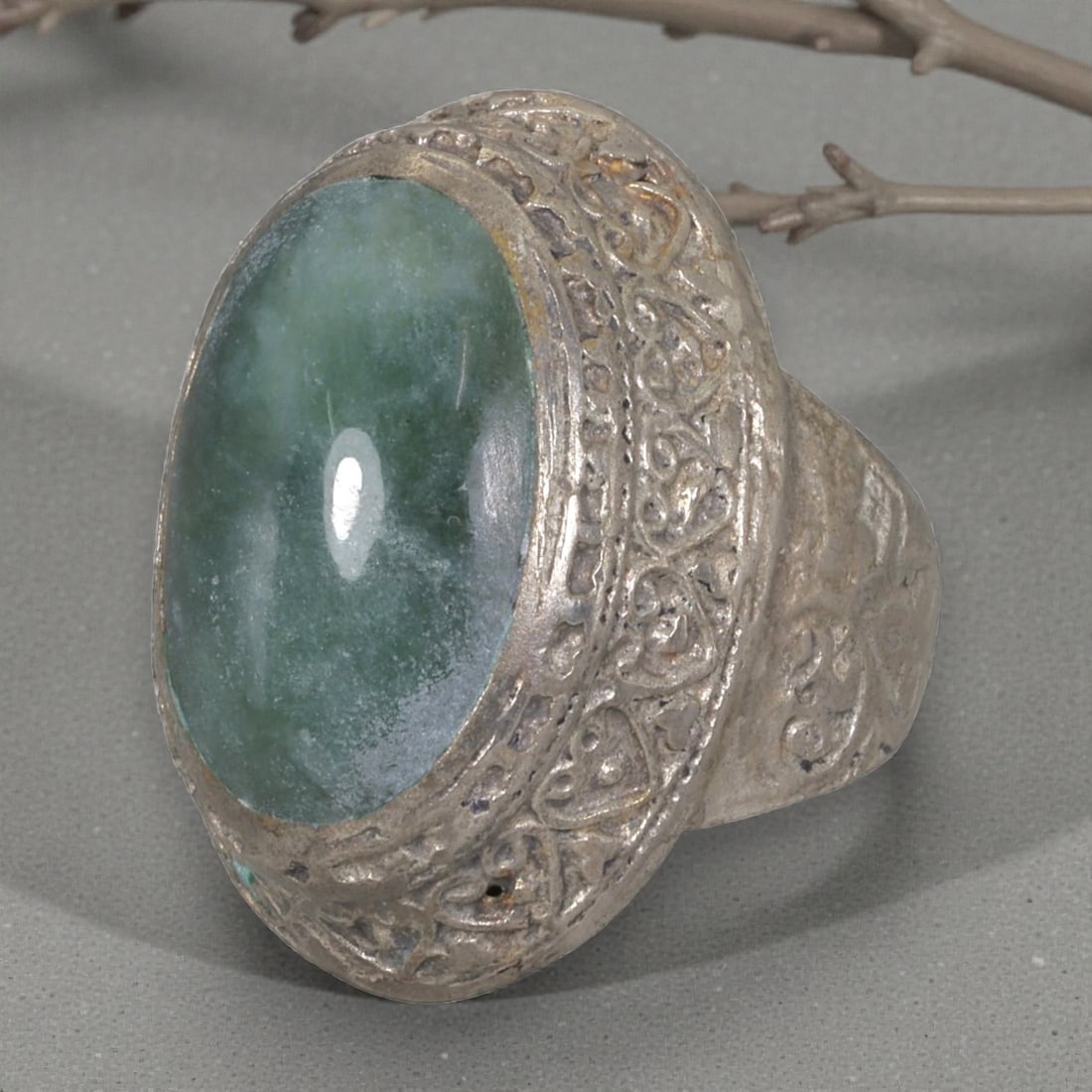A Middle Eastern Silver Plated Green Agate Ring, Late 19th-Early 20th Century, 3AK: The ring is centered with a large oval cabochon green agate stone exhibiting natural mottled green tones, set within a domed bezel featuring intricate engraved foliate motifs. The shoulders are also d