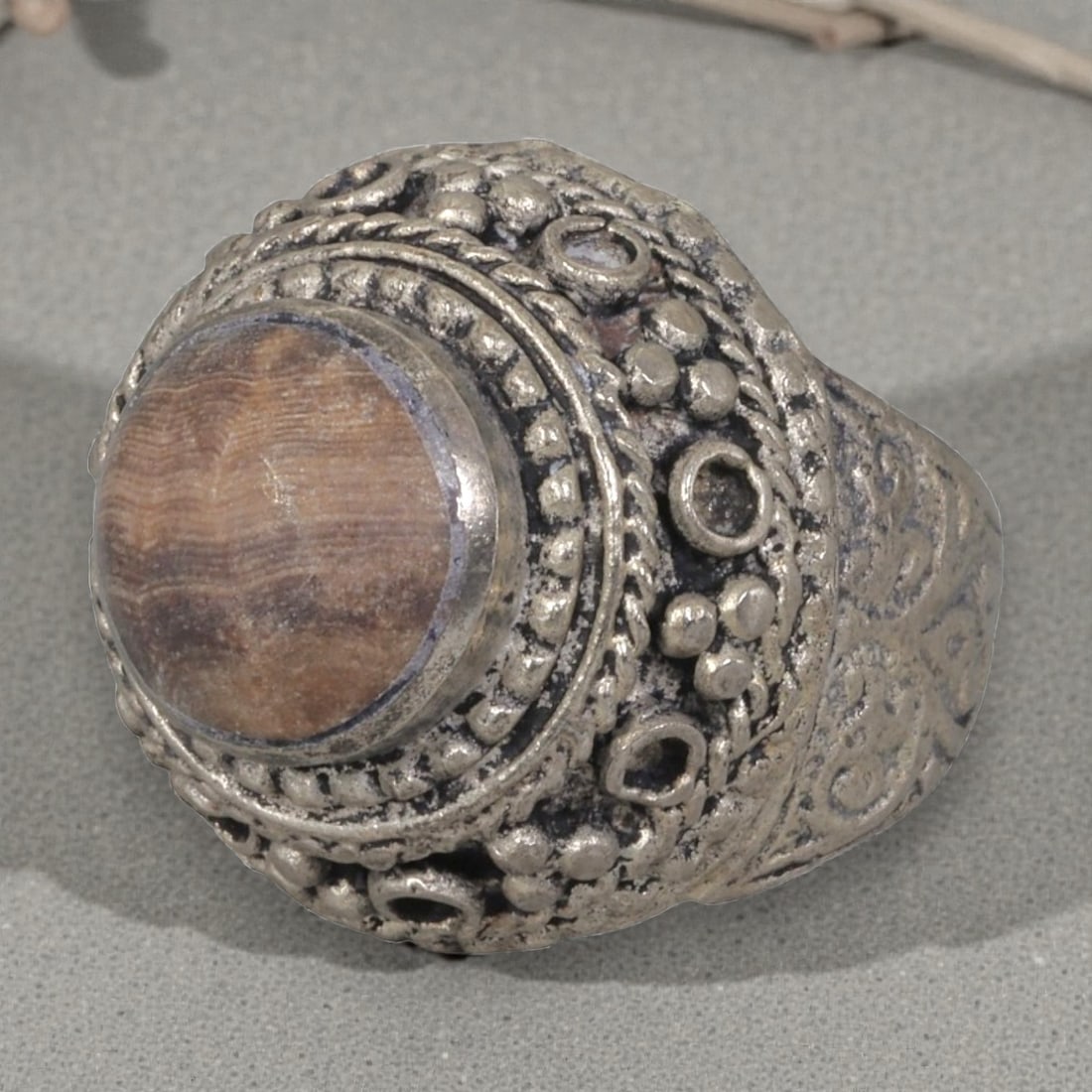 A Middle Eastern Silver Plated Banded Agate Ring, Late 19th-Early 20th Century, 6AK: The ring is centered with a round cabochon banded agate stone exhibiting natural layered brown striations, set within an ornate domed bezel decorated with beaded granulation, twisted wirework, and emp