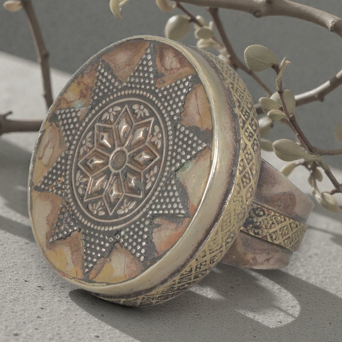 A Central Asian Silver Plated Geometric Star Motif Ring, Late 19th-Early 20th Century, 14AK: Crafted from silver plated metal with brass accents, featuring a wide, open band. The circular bezel displays a multi-layered star motif: a central eight-pointed star surrounded by small floral detail