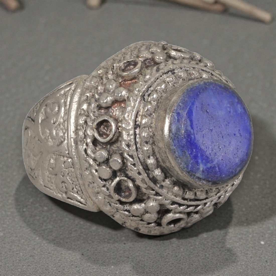 A Middle Eastern Silver Plated Lapis Lazuli Ring, Late 19th-Early 20th Century, 8AK: The ring is centered with a round cabochon lapis lazuli stone exhibiting deep blue color and natural pyrite flecks, set within an ornate domed bezel decorated with beaded granulation, twisted wirework
