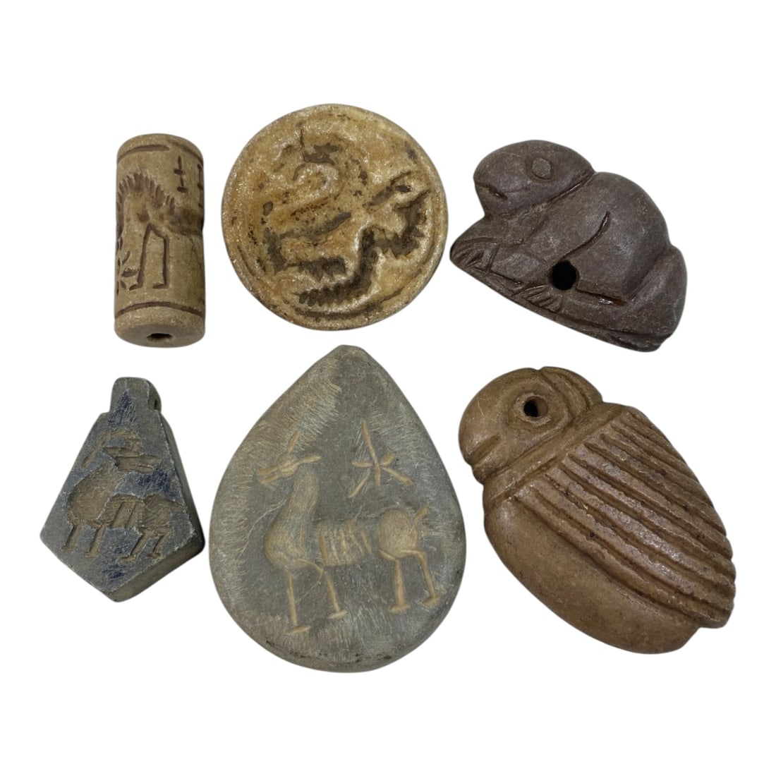 Lot of 6 Ancient Near Eastern Stone Seals and Zoomorphic Amulets, 50AB: The group comprises six carved stone objects, including a cylinder seal, disc seal, teardrop-shaped seal, triangular pendant, and two zoomorphic amulets, crafted from light and dark grey stone, and br