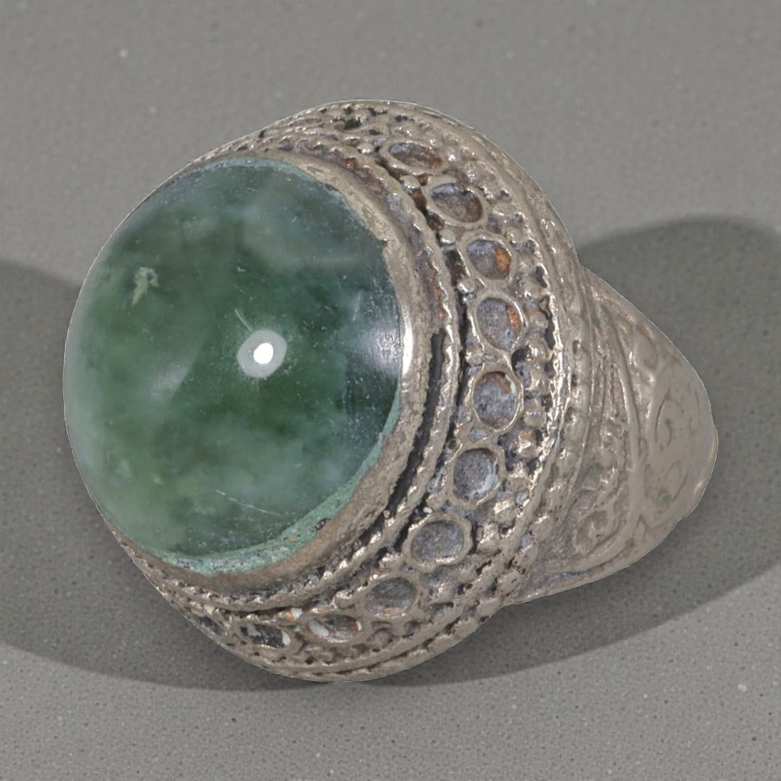 A Middle Eastern Silver Plated Green Agate Ring, Late 19th-Early 20th Century, 13AK: The ring is centered with a large round cabochon green agate stone exhibiting natural mottled green tones, set within an ornate domed bezel featuring openwork circular patterns and beaded granulation.